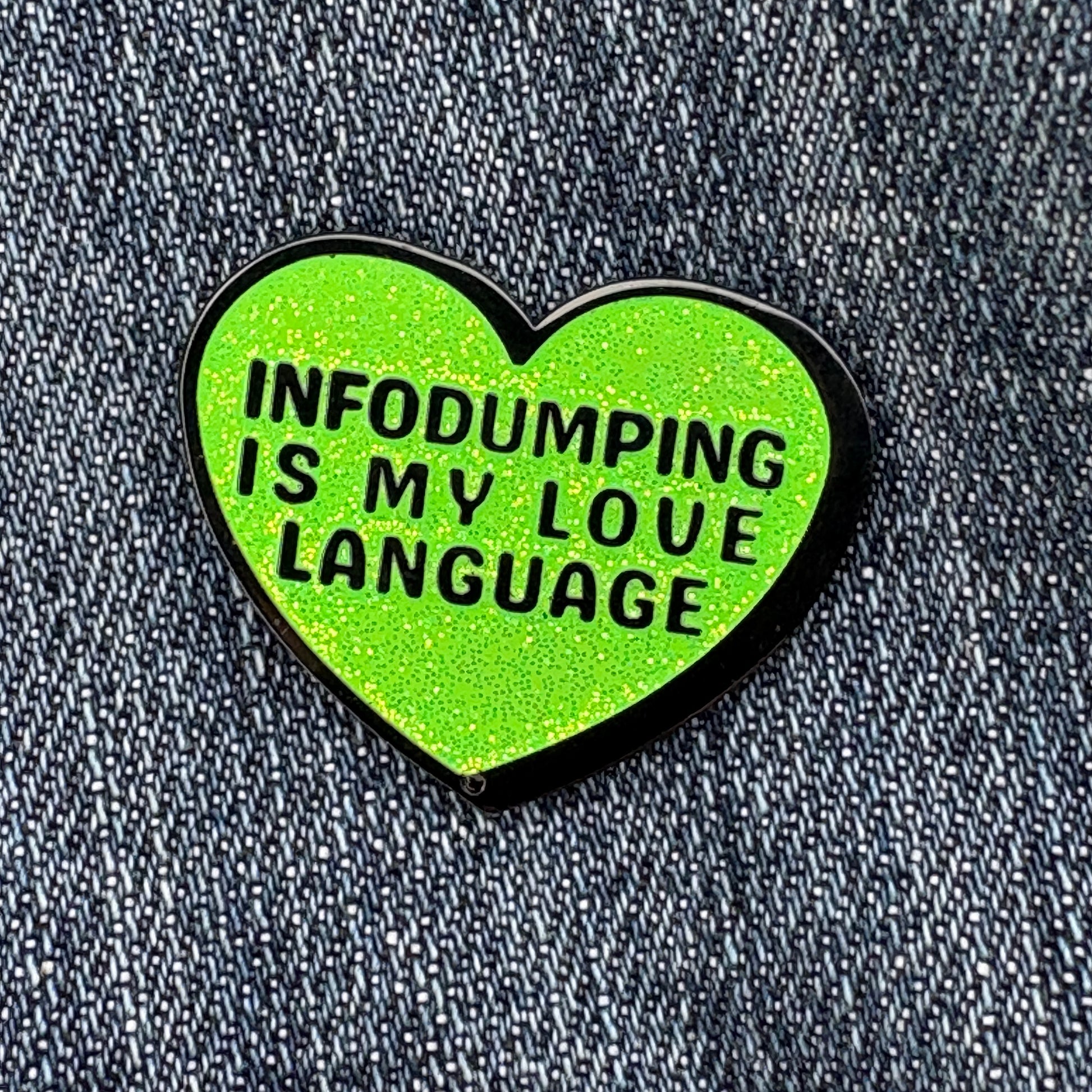 Heart-shaped green enamel pin with text 'INFODUMPING IS MY LOVE LANGUAGE' on a denim background